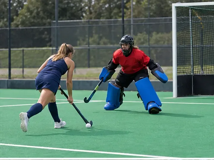 A field hockey attacker dribbles toward a goalkeeper advancing from the goal during a one-on-one shoot-out