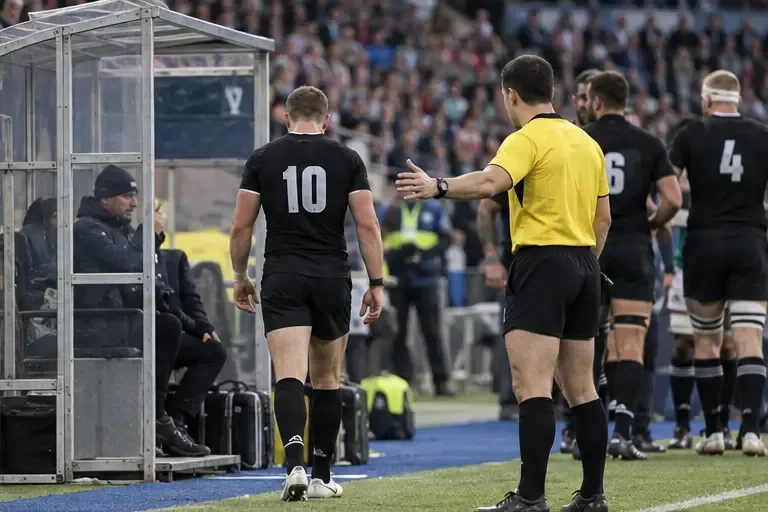 A rugby player walks toward the sideline as the referee gestures toward the sin-bin area