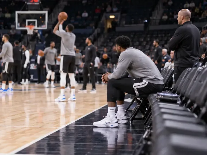 Basketball players warm up on court while one player sits with staff near the sideline.