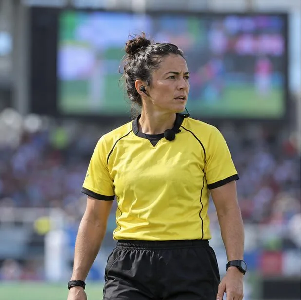 A rugby referee stands on the field with a stadium display blurred behind her, evoking the first check of a pool standings table.