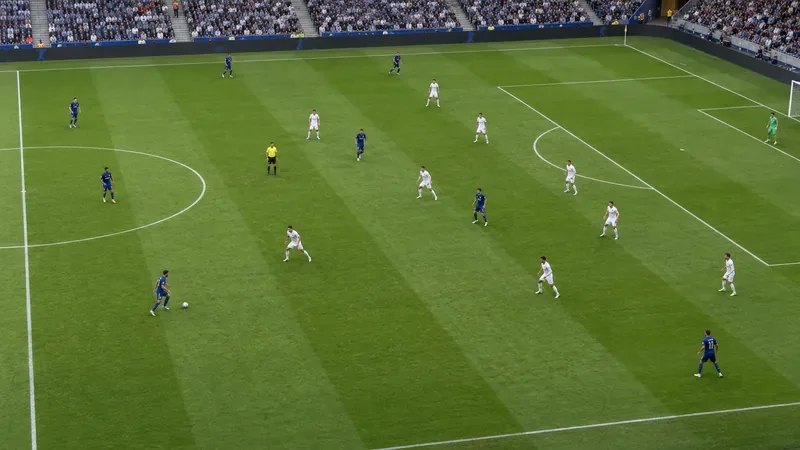 Een overzichtsfoto van spelers in blauwe shirts verspreid over het veld, terwijl verdedigers in witte shirts een compacte formatie vormen.
