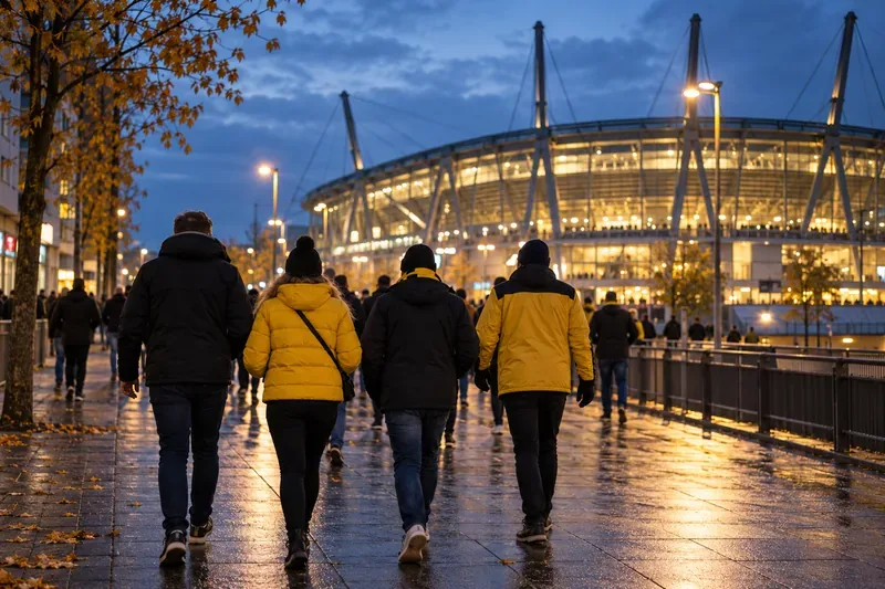 Supporters in gele en donkere jassen lopen in de schemering richting een fel verlicht stadion.