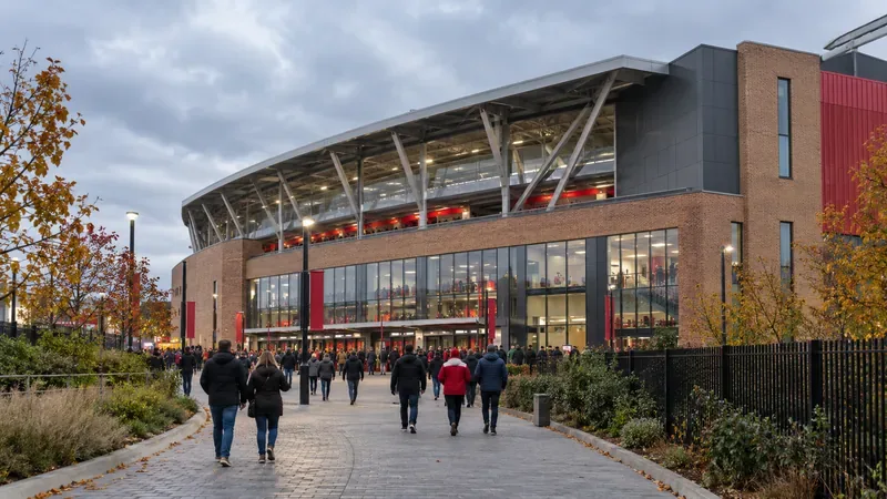 Fans lopen op een bewolkte herfstmiddag richting een compact, modern voetbalstadion van baksteen en glas in West-Londen.