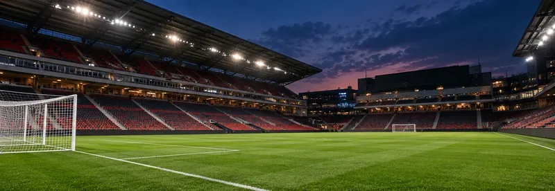 Een leeg, verlicht voetbalstadion met rode en zwarte tribunes en een doel onder een avondhemel.