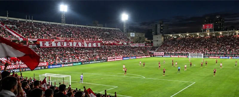 Tribune biancorosse, luci dello stadio e un campo da calcio visto da una tribuna gremita di gente.