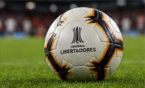 Un pallone da calcio bianco con pannelli grafici neri, oro e arancioni, adagiato su un prato verde, sullo sfondo sfocato di uno stadio.