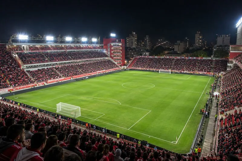 Una panoramica di un campo da calcio verde e delle tribune rosse gremite, illuminate dai riflettori in una notte cittadina.