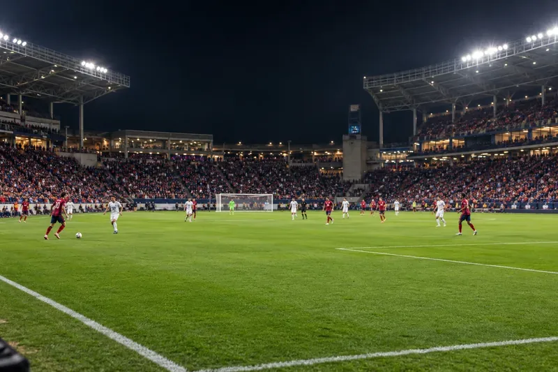 Een breed overzicht van een voetbalstadion 's nachts, met het veld, het doel, de spelers en de verlichte tribunes.