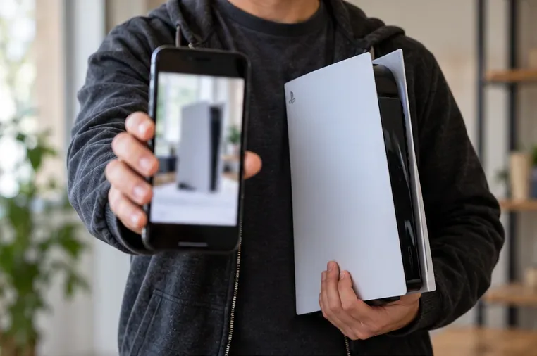 A seller holds a PlayStation 5 next to a phone showing a fresh photo of the same console.