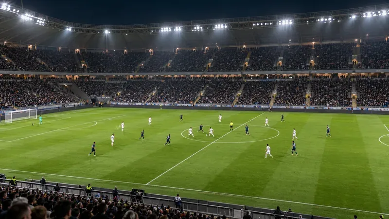 Una vista panorámica de un partido de fútbol nocturno en un campo verde bajo los focos del estadio, con las gradas llenas de espectadores.
