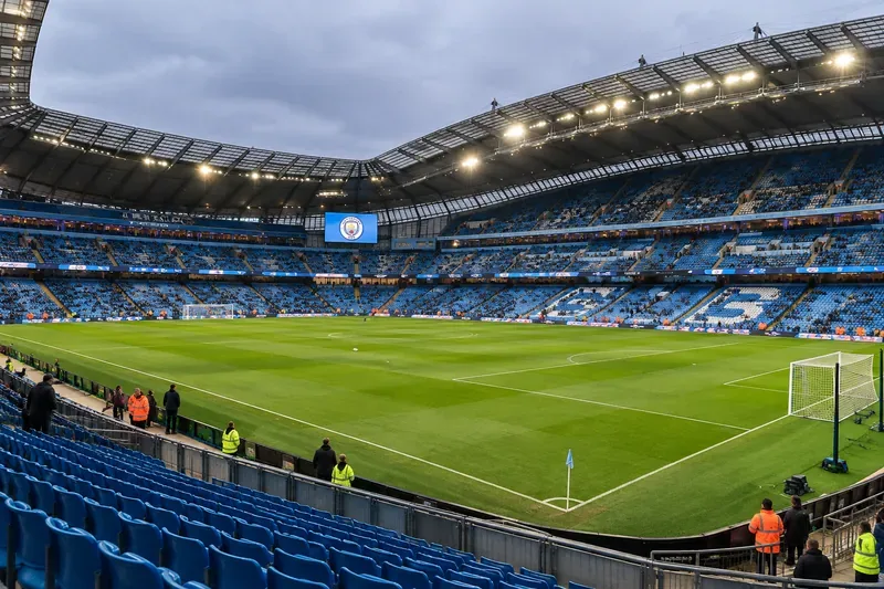 Een panoramisch uitzicht op een groen voetbalveld, een doel, een hoekvlag en blauwe tribunes onder de verlichte dakbalken van het stadion, vlak voor een wedstrijd.
