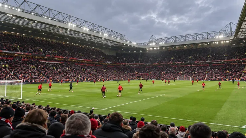 Footballers in red warm up on a green pitch inside a full stadium beneath an overcast sky.