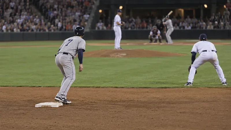 A baserunner stands on second base and takes a short lead while a fielder waits several steps away.