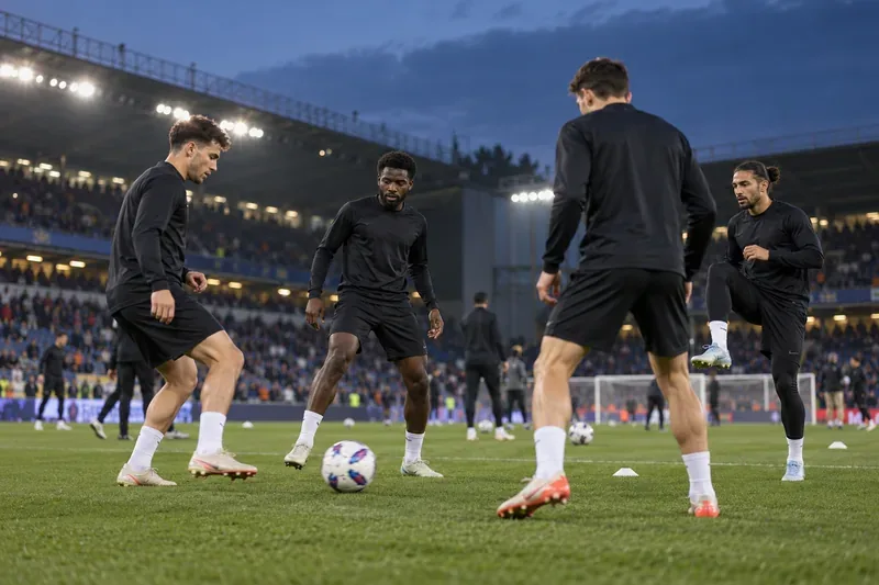 Quattro calciatori in semplici tute da allenamento scure si riscaldano passandosi un pallone su un campo illuminato.