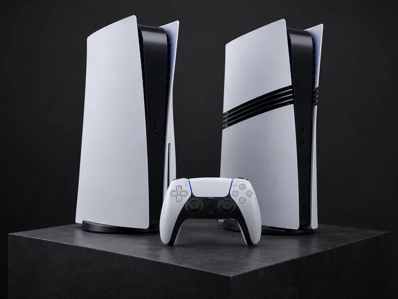 Um PS5 branco e preto ao lado do PS5 Pro, cujos painéis laterais brancos são divididos por três faixas pretas, com um controle DualSense.