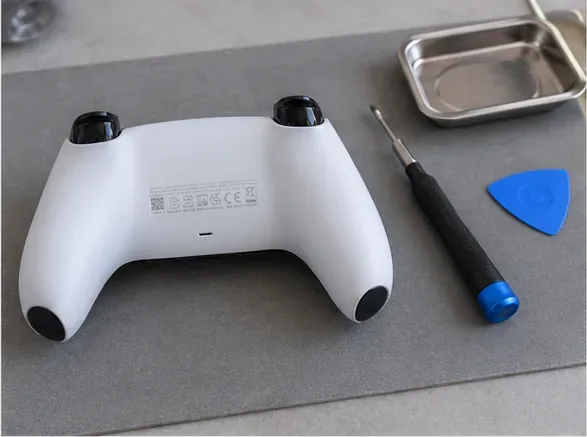 Controle DualSense desligado ao lado de uma chave de fenda Phillips, uma palheta de plástico para abertura e uma bandeja de peças.