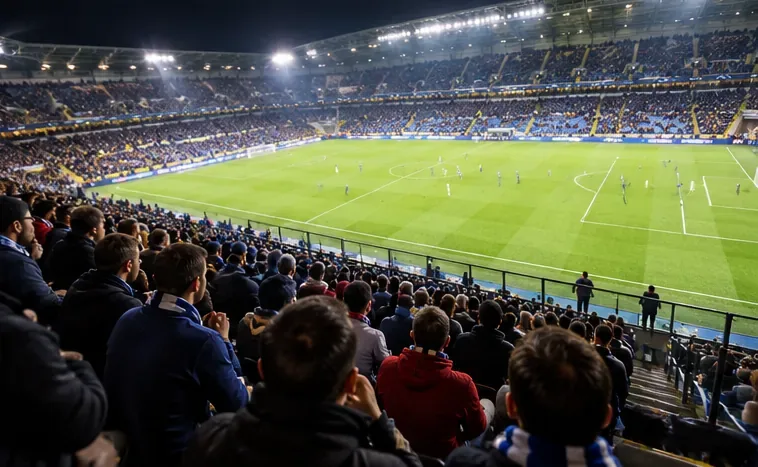 Toeschouwers vanaf een verhoogde tribune met uitzicht op een fel verlicht voetbalveld en spelers in de verte.