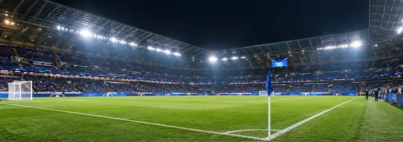 Een verlicht voetbalstadion met een groen veld, een blauwe hoekvlag en toeschouwers op overdekte tribunes.