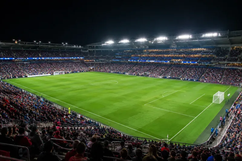 Breedbeeld van een verlicht voetbalstadion met een groen veld en fans op volle tribunes.