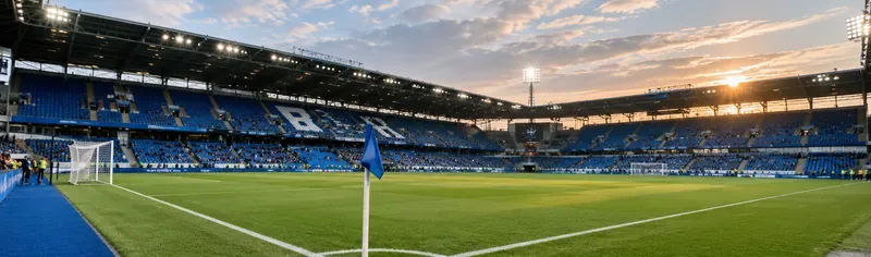 Een verlicht voetbalstadion met blauwe tribunes, een groen veld en een blauwe hoekvlag bij zonsondergang.