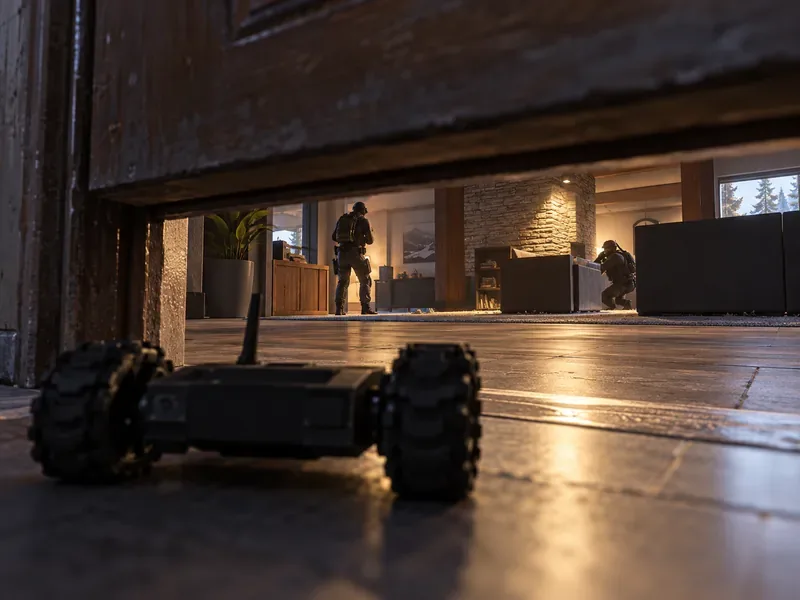 A small wheeled scouting drone looks beneath a doorway toward two distant defenders in a room.