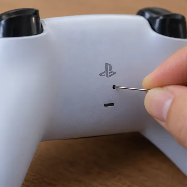 A fine-tipped pin is pressed into the small reset-button hole on the back of a DualSense-style controller.
